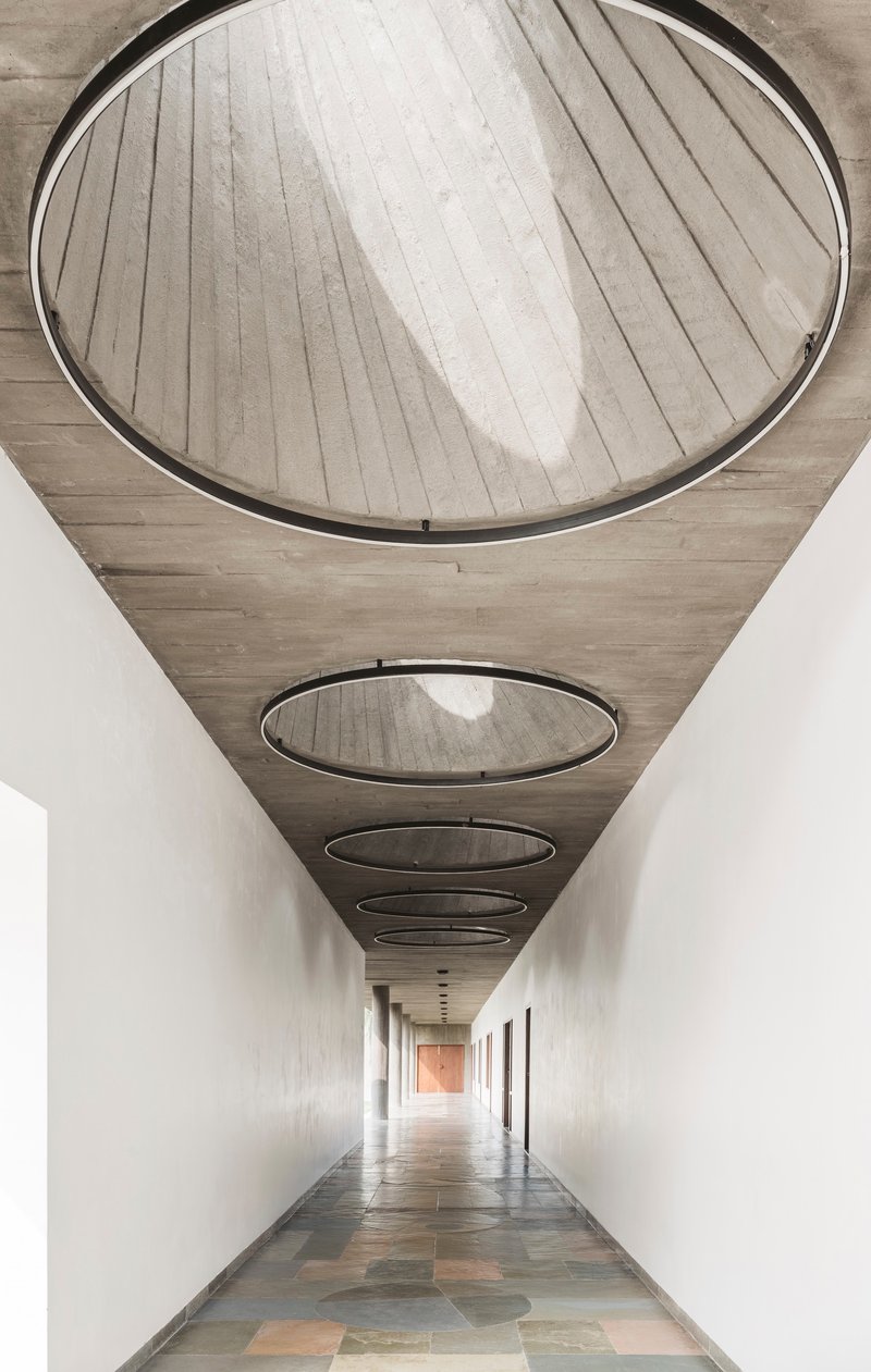 Corridor with circular skylights set in board-formed concrete ceiling above polished multicolored stone floor