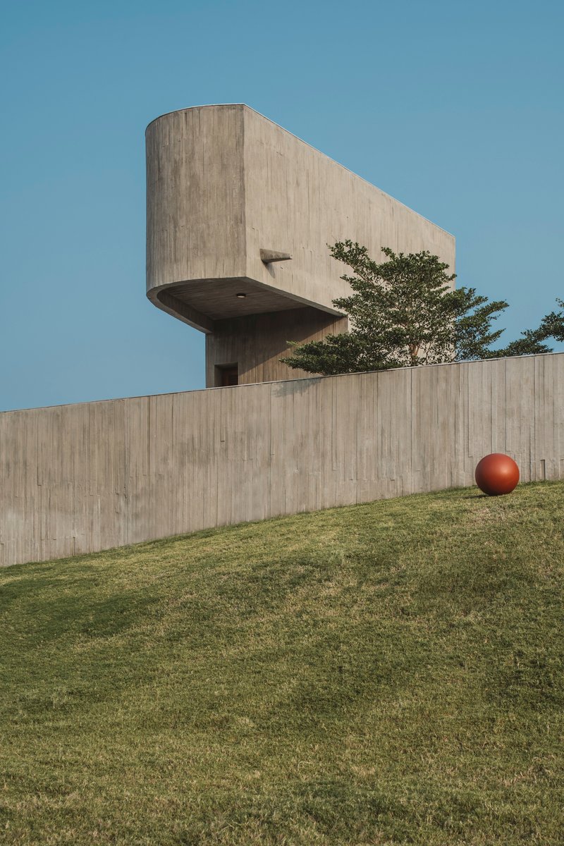 Board-formed concrete tower cantilevering over a curved retaining wall with a sphere on the grass