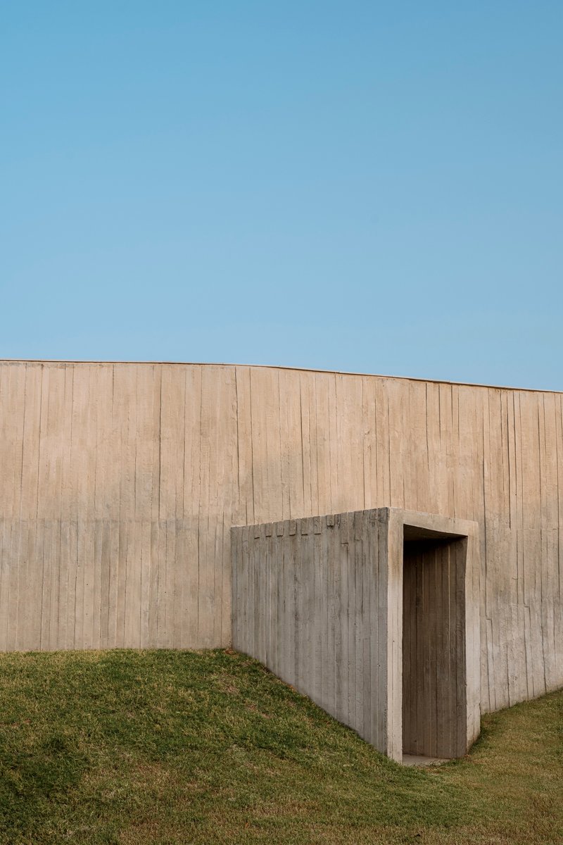 Board-formed concrete wall with recessed doorway threshold against a grass slope under clear sky