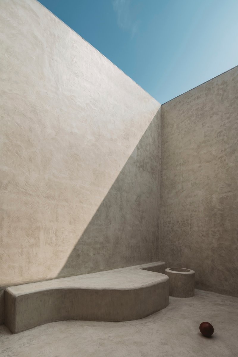 Concrete courtyard with angled walls, sculpted bench, cylindrical planter, and sharp diagonal shadows under open sky