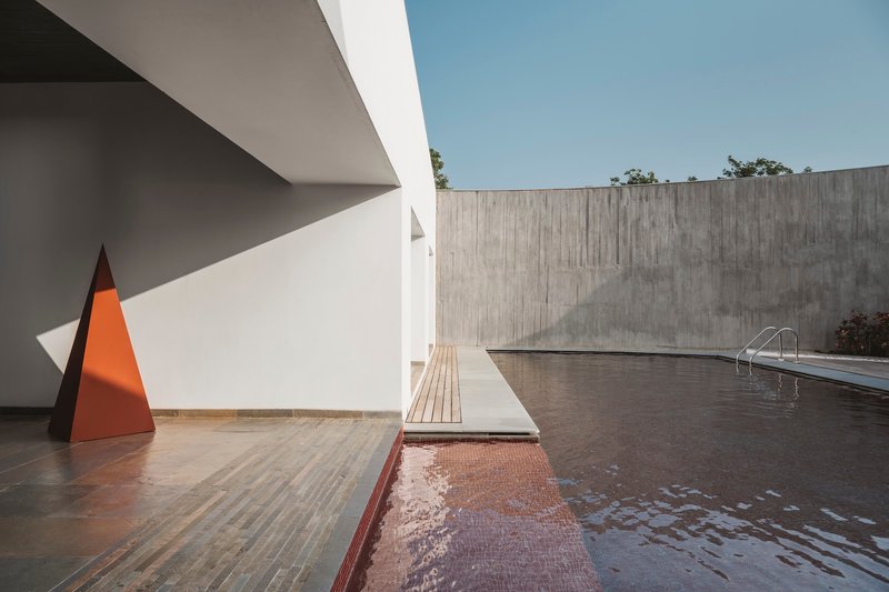 Pool terrace with red tile basin and white deck alongside concrete wall and orange pyramid sculpture