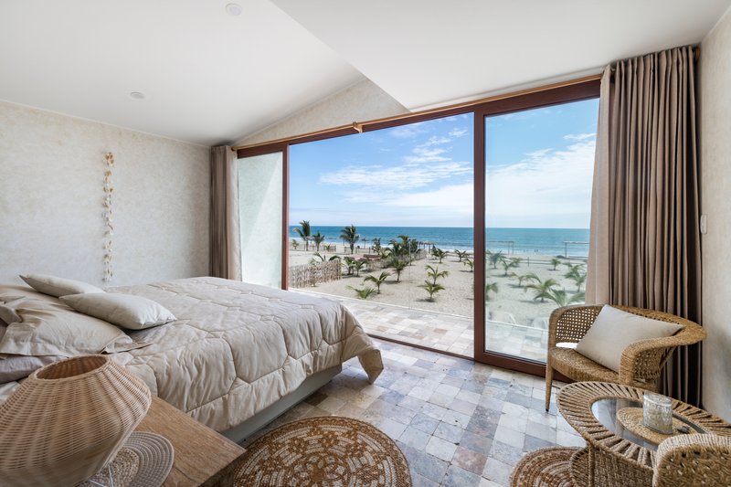 Bedroom with floor-to-ceiling glazing framing ocean views and young palm trees in the sand