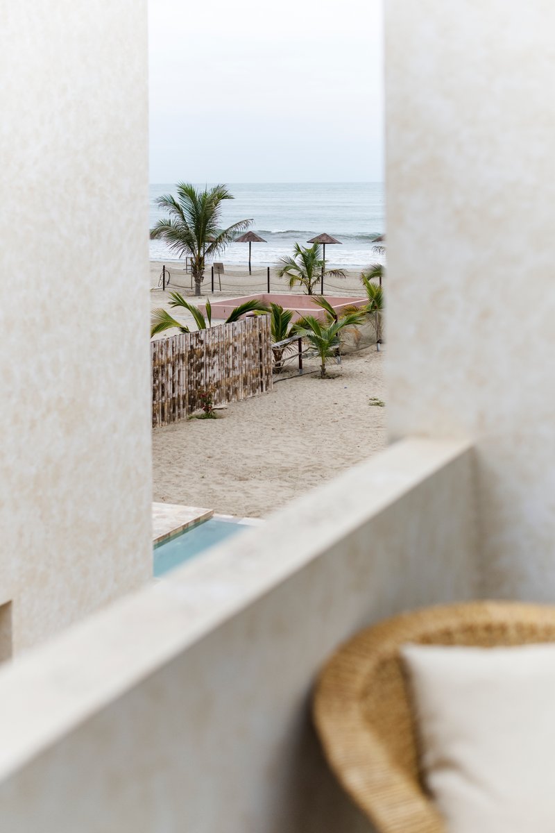 View through narrow opening between white walls toward the beach with palms and thatch umbrellas