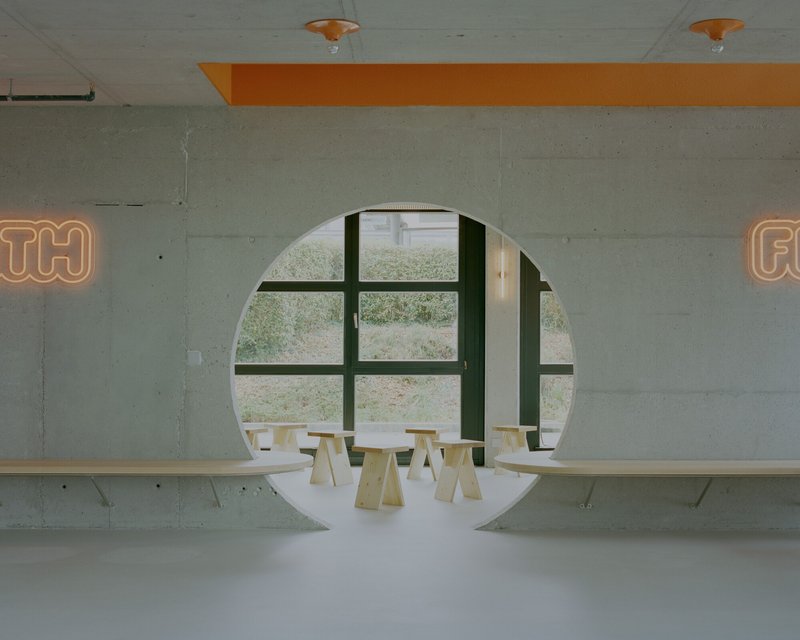 Circular portal framing a window mullion and plywood stools beneath orange neon lettering on concrete