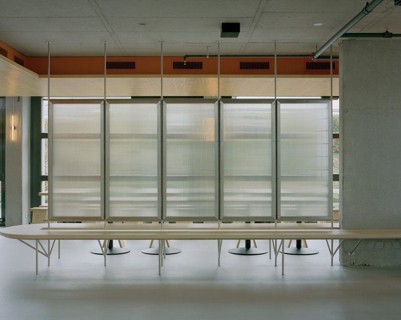 Translucent corrugated wall panels with horizontal orange banding above a long plywood table and green columns