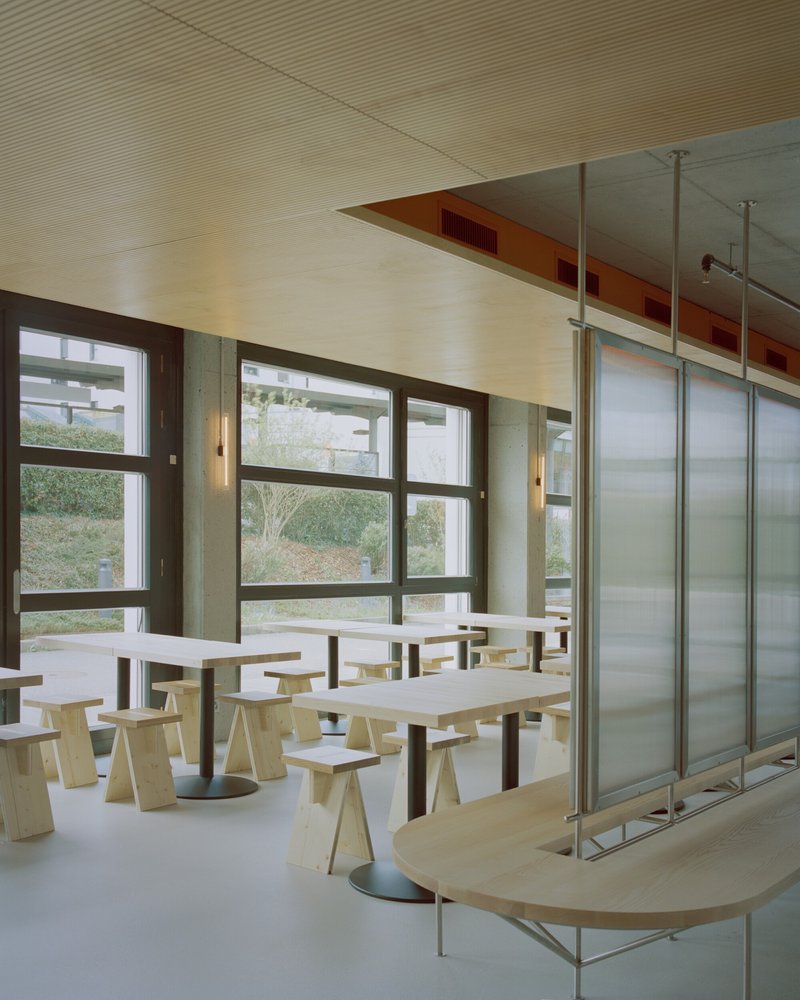 Dining hall with pale wood tables and stools beneath a ribbed plywood ceiling and translucent glass partitions