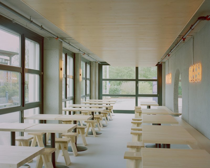 Row of light wood tables and benches along a glazed wall with exposed conduit under overcast daylight
