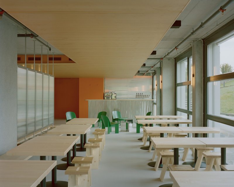 Dining hall with exposed plywood ceiling, translucent wall panels, and green plastic chairs near tall windows