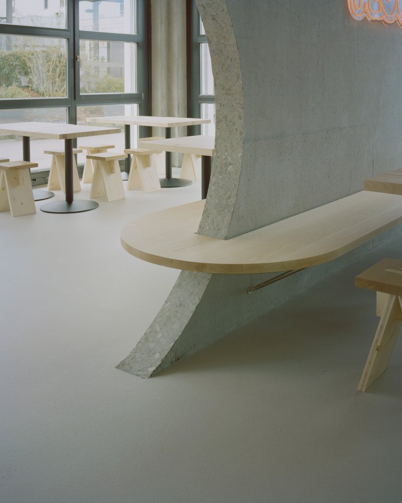 Light wood bench with angled concrete support beside plywood seating and tables near a window