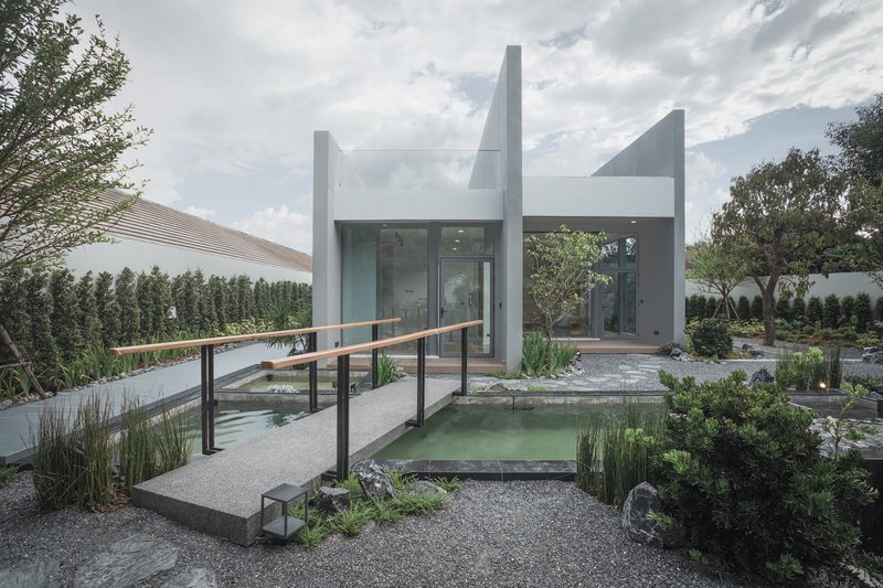 Gravel courtyard with reflecting pond and timber bridge leading to white plastered volumes under clouded skies