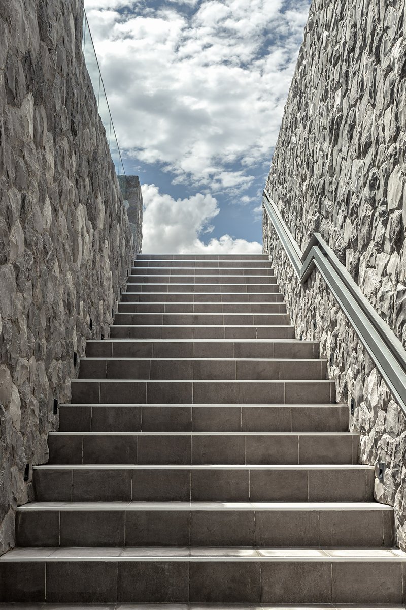 Tiled staircase between stone walls ascending toward a cloudy sky with metal handrails