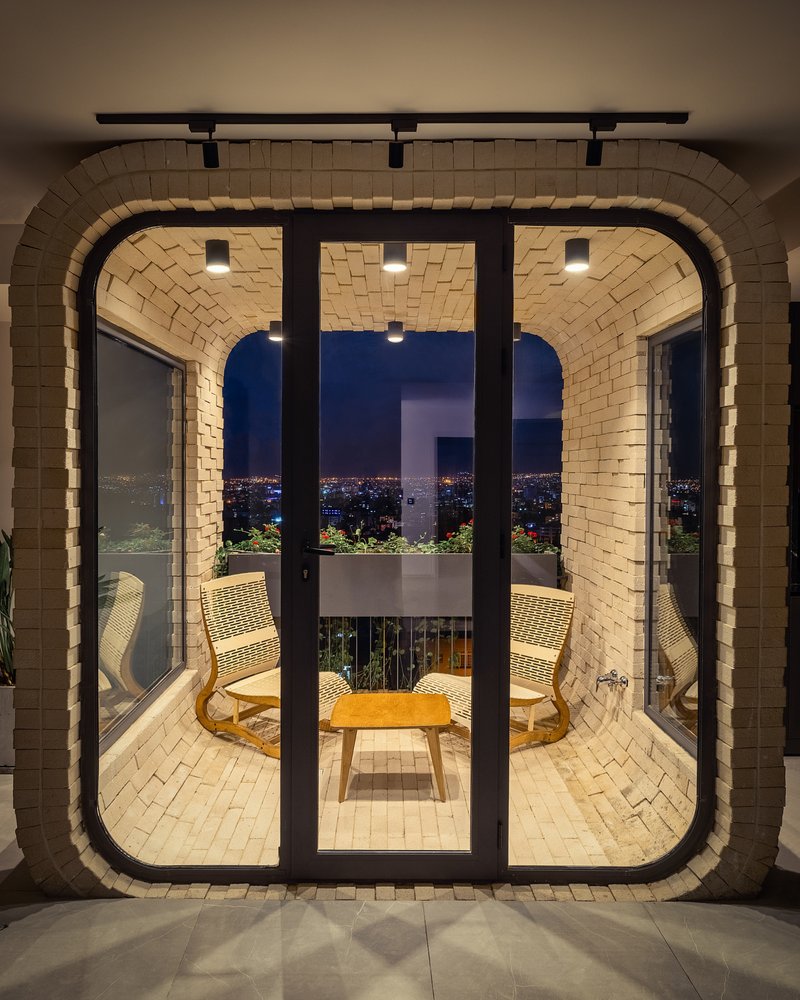 Vaulted brick pavilion with black-framed glass doors opening to a balcony overlooking city lights at dusk