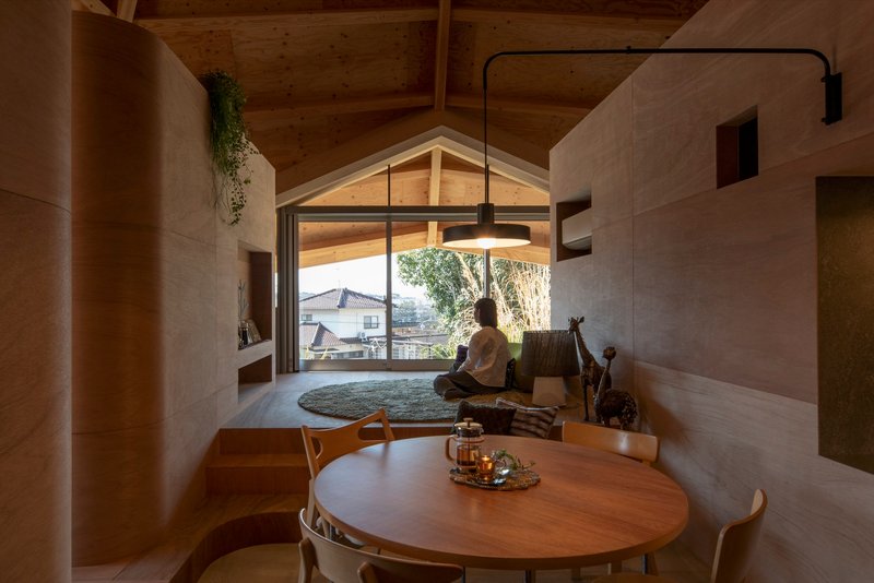Living area with plywood partitions and round dining table where a person sits on a rug near the gabled window
