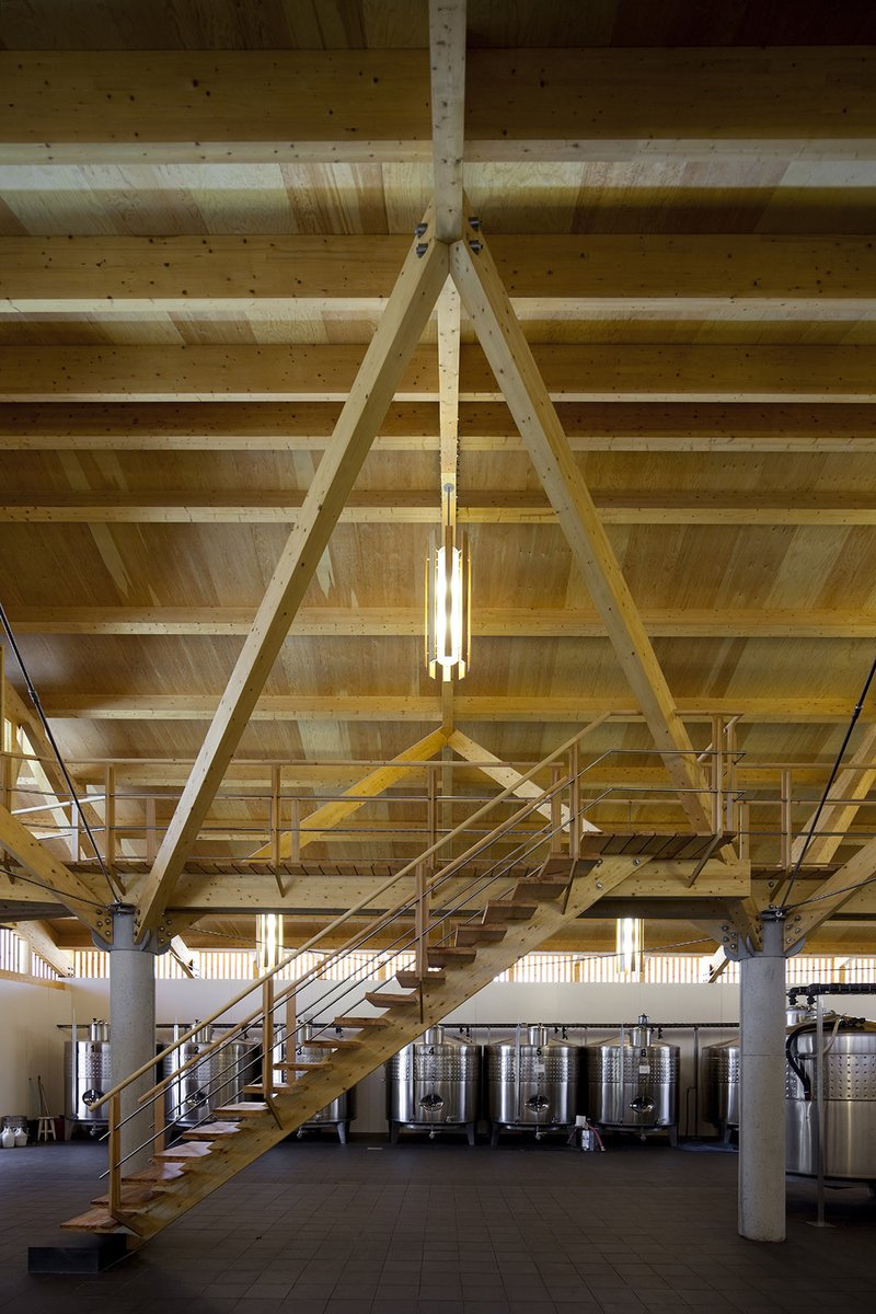 Interior view of exposed timber A-frame trusses and beams above an open staircase and stainless fermentation tanks