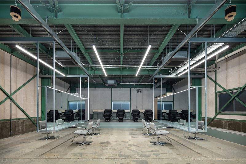 Interior salon with exposed green steel trusses and linear fluorescent lighting above white styling chairs