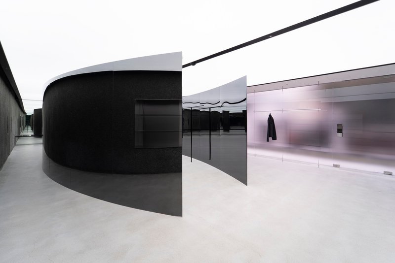 Curved black metal volume alongside polished metal panels with pale resin flooring under diffused daylight