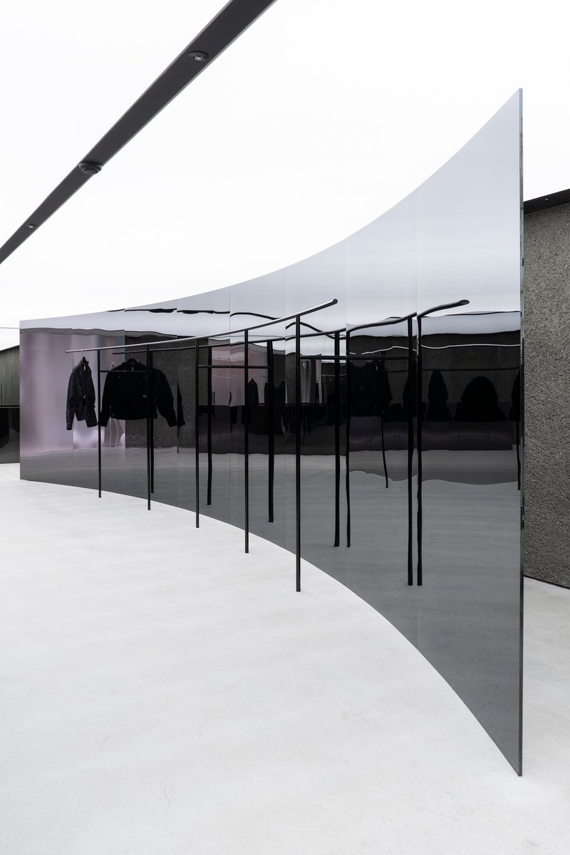 Angled view of clothing display racks behind dark glass with textured stone wall under white fabric ceiling