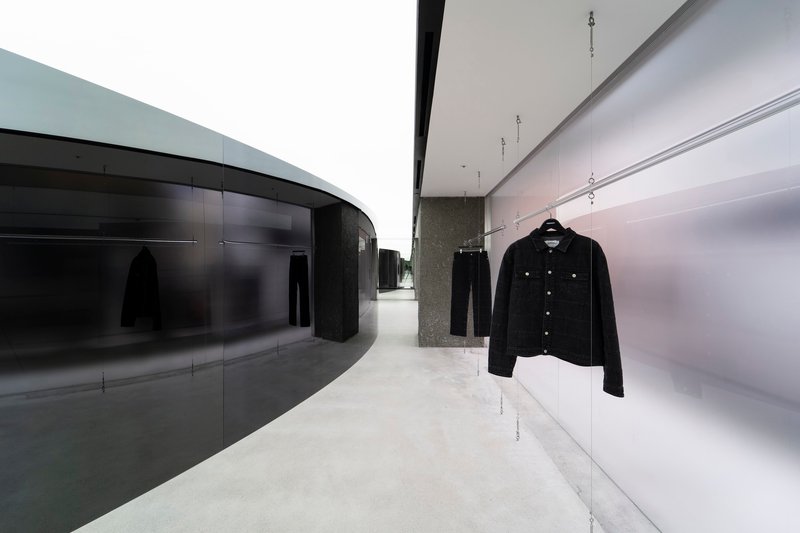 Display hallway with curved dark glass wall opposite illuminated white shelving and hanging denim jacket