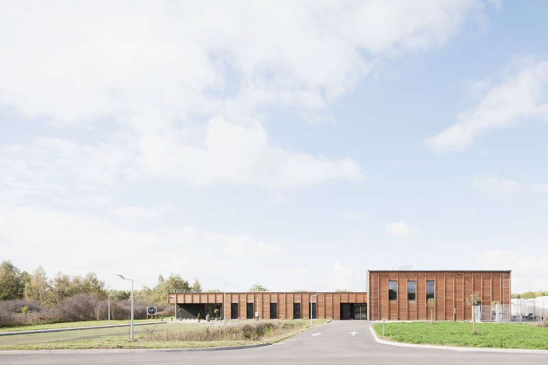 Long horizontal timber-clad building with rhythmic window openings set in a grassy field under cloudy skies