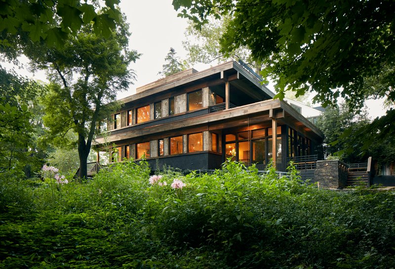 Two-story residence with exposed timber beams and dark cladding framed by lush garden vegetation and mature trees