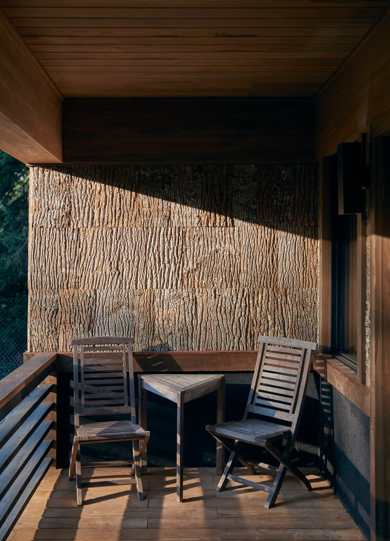 Covered balcony with textured bark-clad wall, timber ceiling, and two chairs catching angled afternoon sunlight