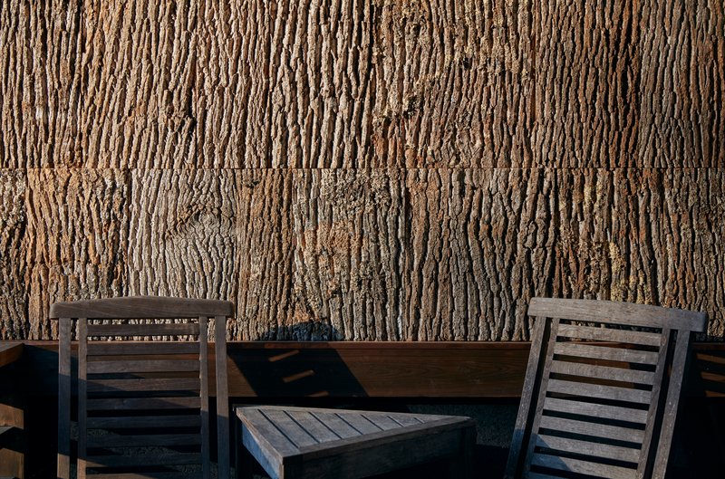 Detail of rough bark wall cladding with two timber chairs casting diagonal shadows on the deck