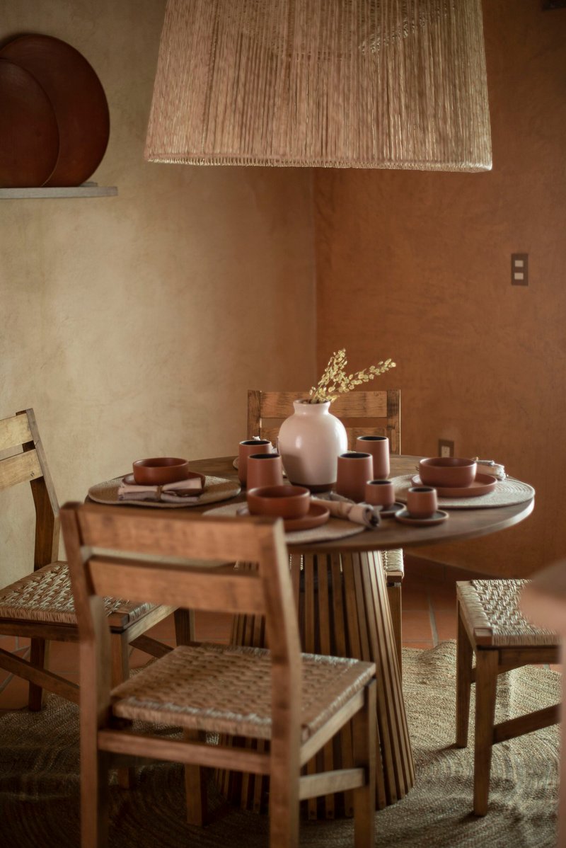 Round timber dining table with woven raffia pendant light and earthenware vessels in warm daylight