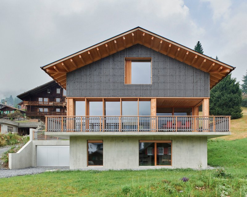 Gabled facade with vertical metal cladding and exposed timber eaves above a concrete base and grass slope