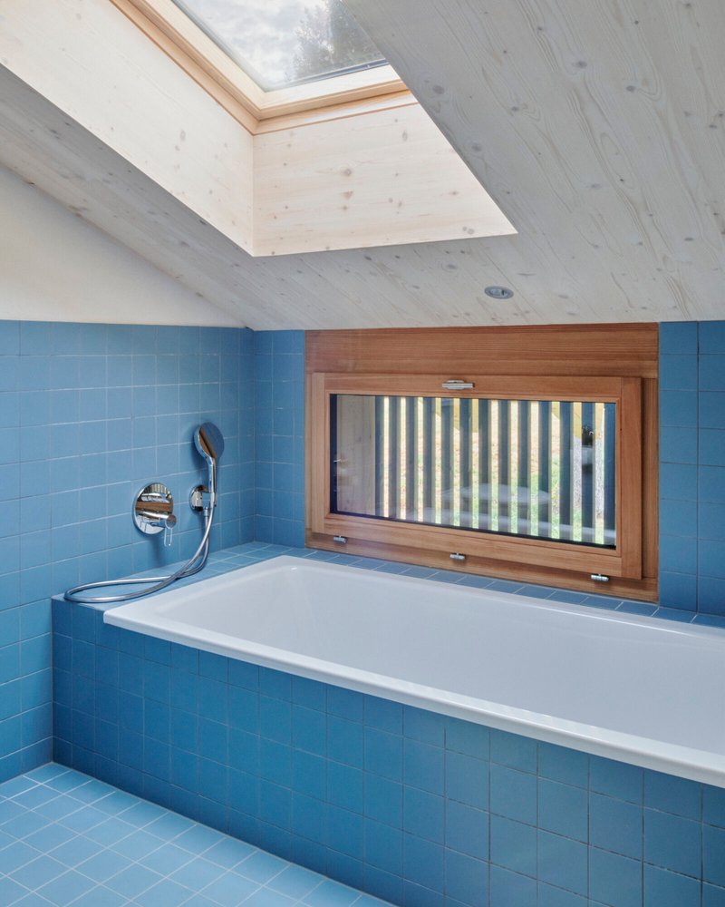 Bathroom with blue tile surround, white bathtub, timber-framed window and skylight above exposed wood ceiling