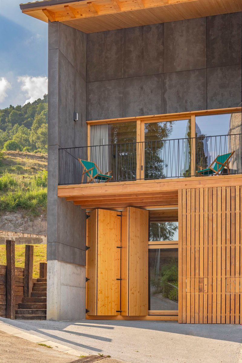 Two-story facade with concrete upper level, timber lower cladding and balcony with two chairs at dusk