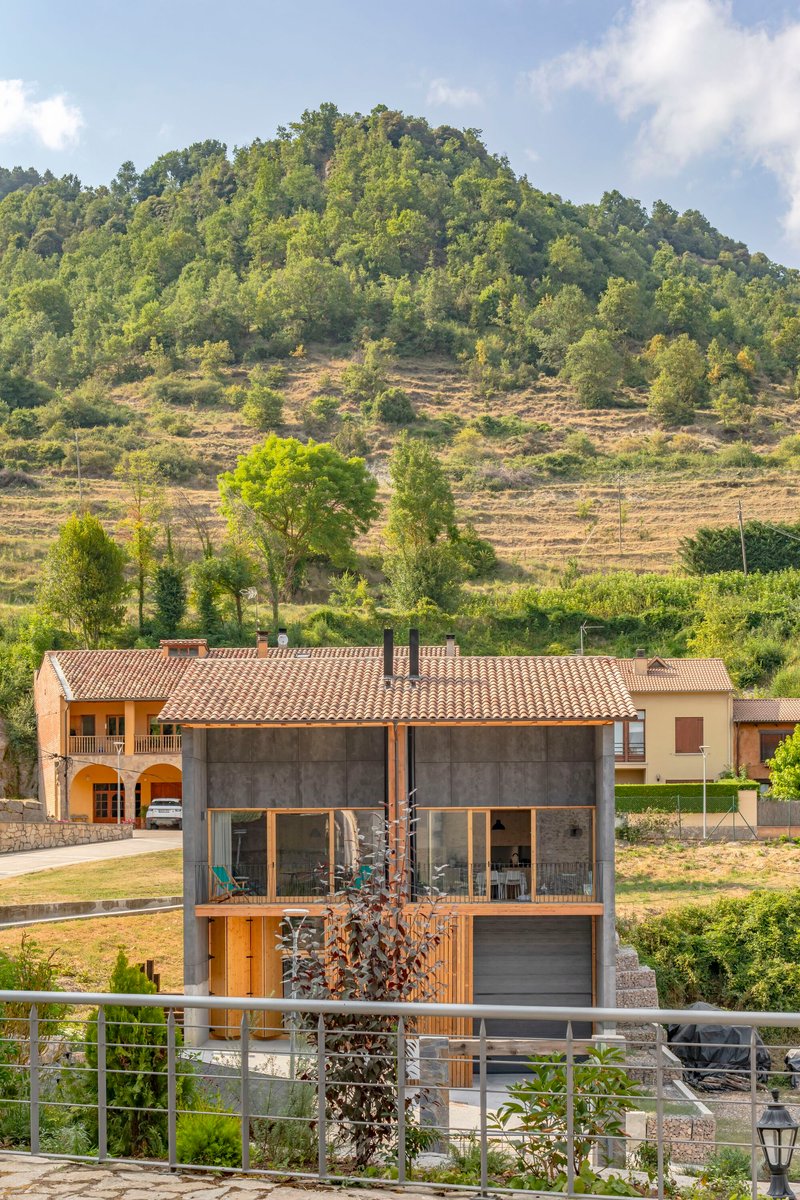 Concrete and timber house with exposed rafters beneath a terraced hillside planted with trees