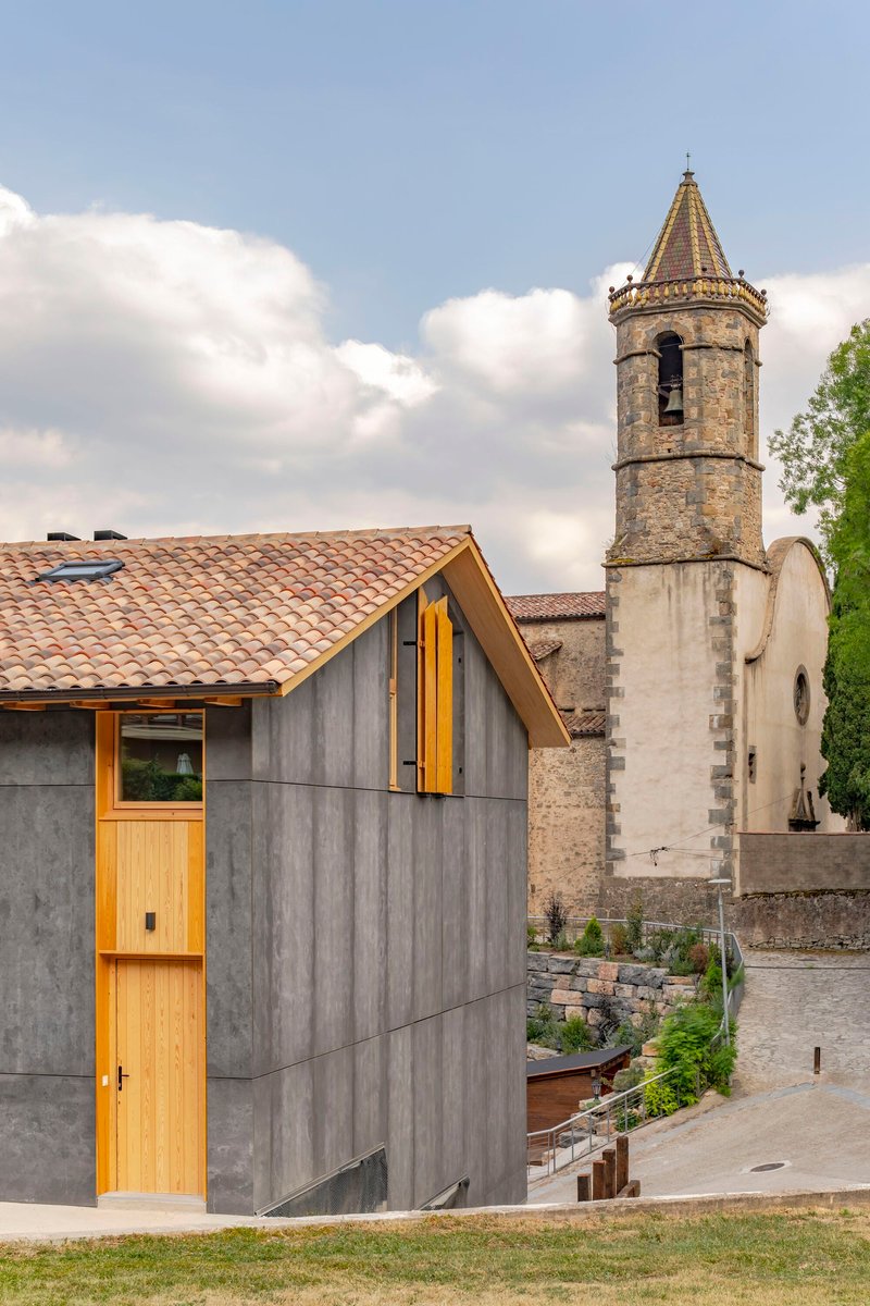 Board-formed concrete facade with timber-framed windows beside a stone bell tower under cloudy skies