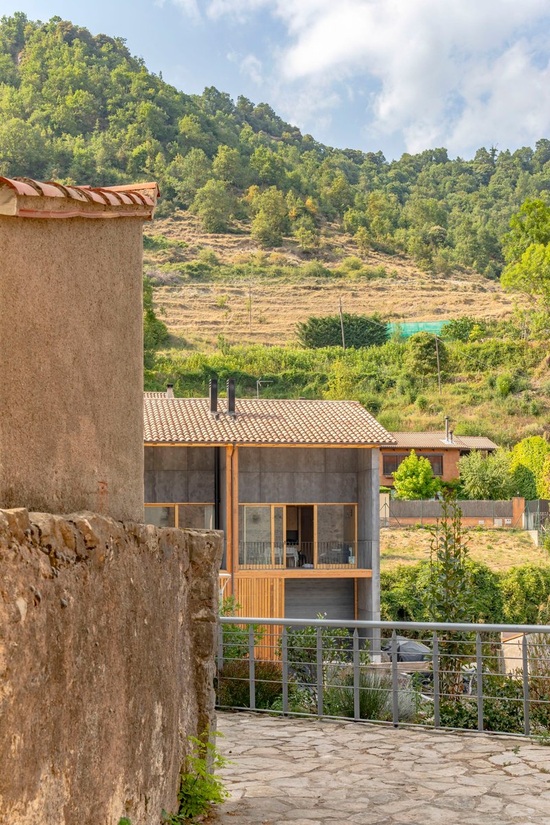 View of the terraced roof and concrete facade nestled among forested hills and surrounding village buildings