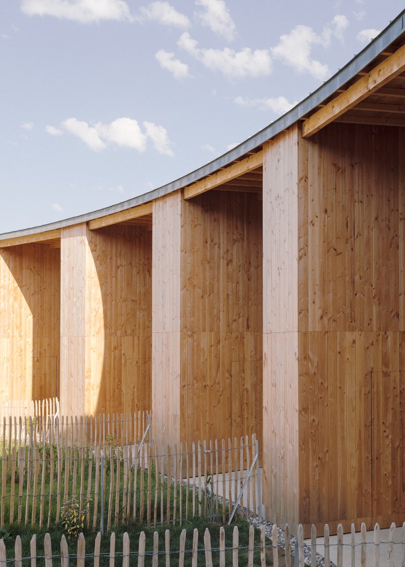 Close-up of vertical timber cladding with chamfered pilasters and white picket fence under a curving metal roof