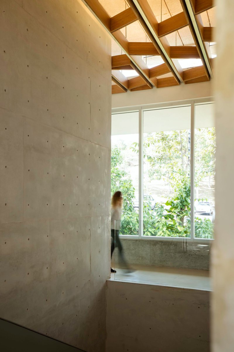 Interior view of concrete wall beside tall window with blurred figure passing through skylit timber-beamed space