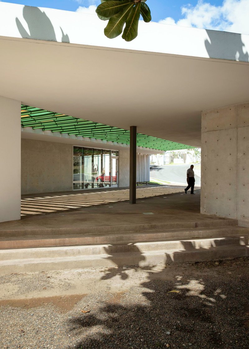 Covered concrete terrace with green slatted ceiling, shadow patterns on floor, and single figure walking through
