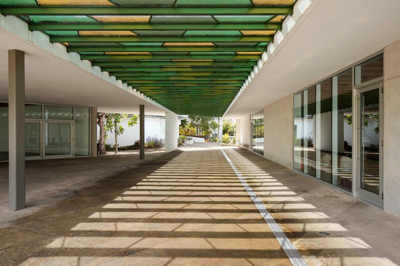 Covered walkway with green corrugated ceiling panels casting striped shadows on concrete pavement