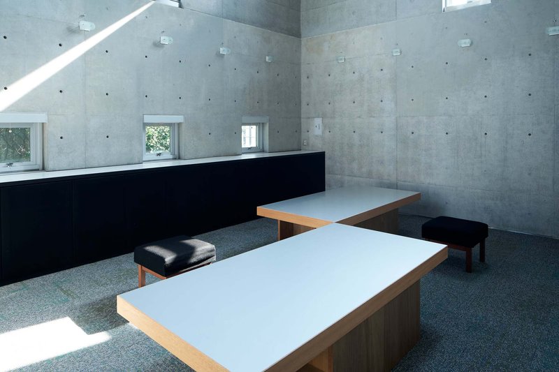 Board-formed concrete interior with low tables and cushioned stools on carpet under sloped skylights
