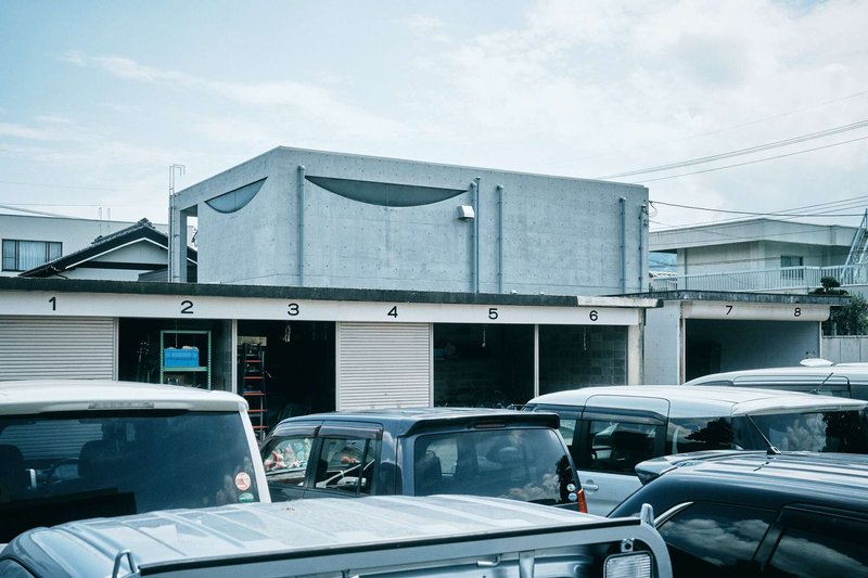 Street view of concrete volume above numbered garage doors with parked vehicles in foreground
