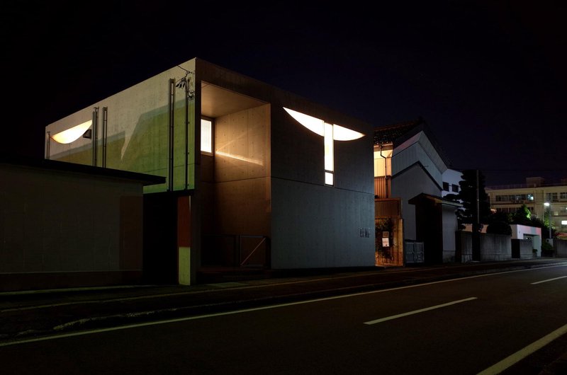 Street view of the illuminated upper volume with curved skylights at night beside neighboring houses