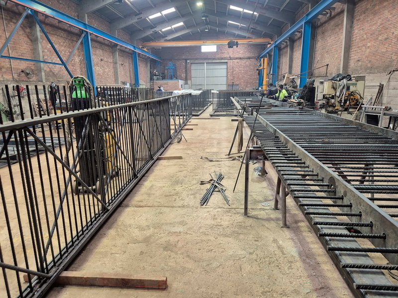 Fabrication workshop with rebar assemblies and a worker in high-visibility vest under blue steel trusses
