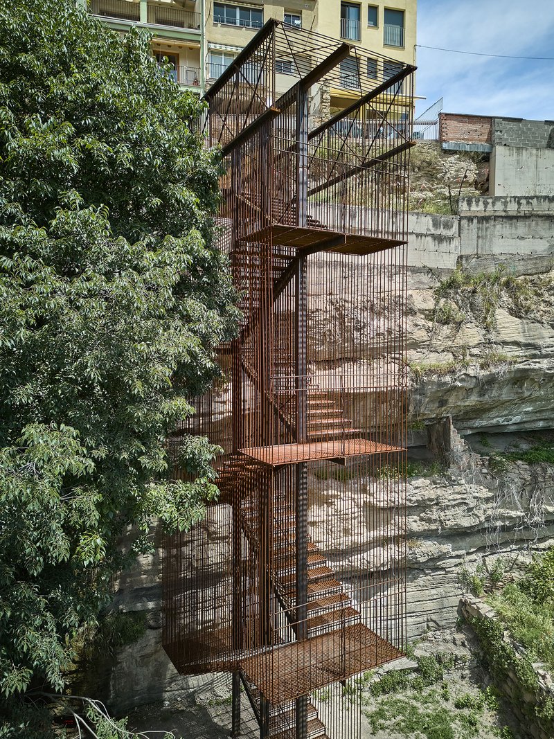 Frontal view of the perforated steel stair tower emerging from layered sedimentary rock beside mature trees