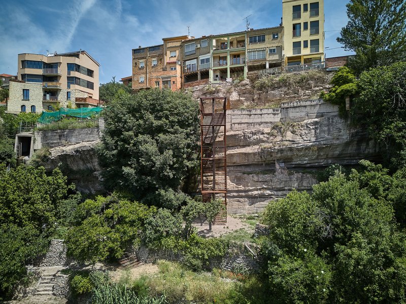 Metal staircase descending a weathered stone cliff face beneath colorful residential buildings and summer foliage