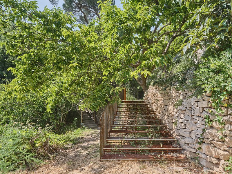 Metal frame stair structure under leafy tree canopy beside a dry-stacked stone retaining wall