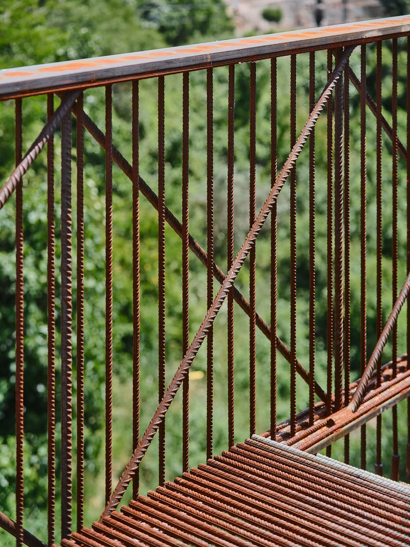 Exposed rebar railing with diagonal cross-bracing against a backdrop of green foliage