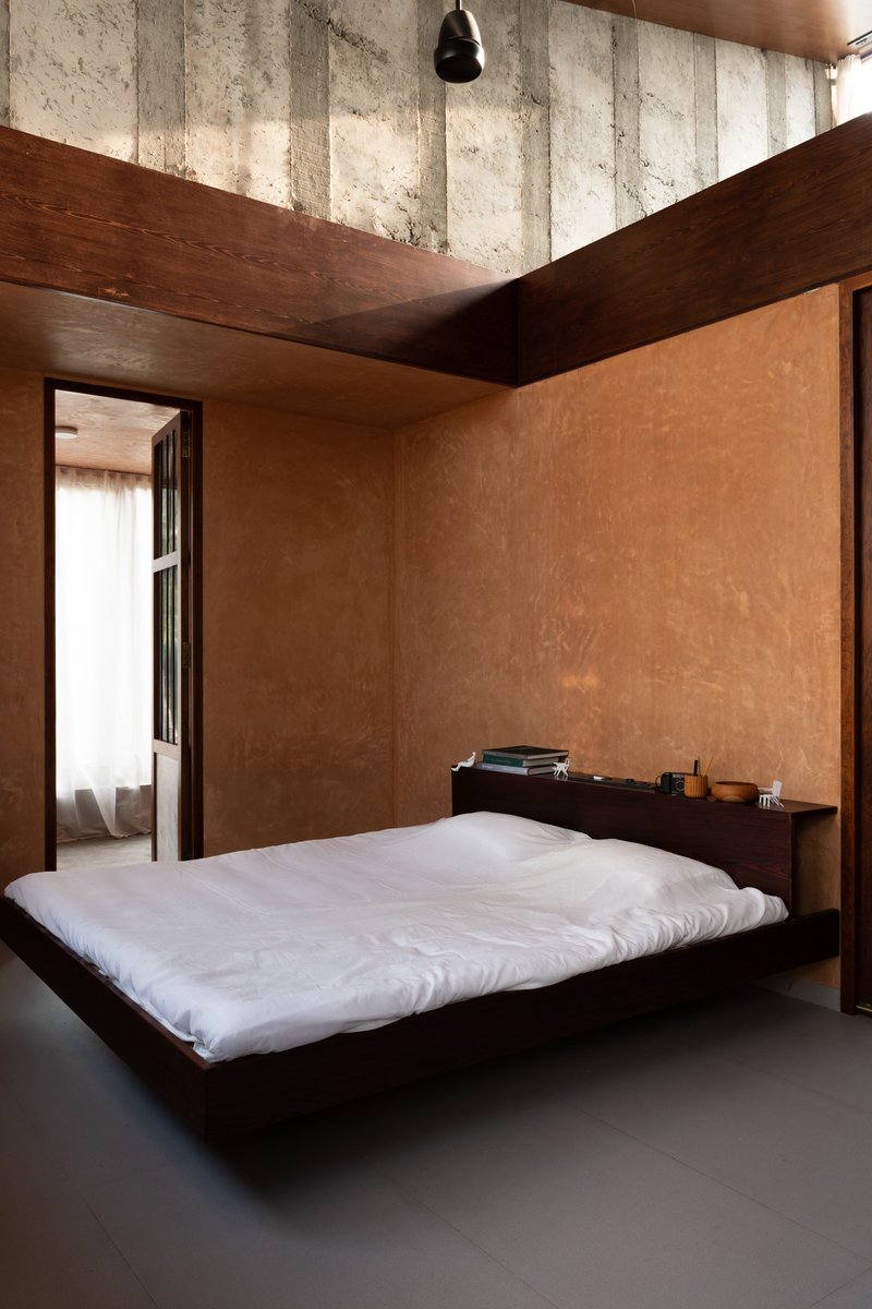 Sleeping space with low platform bed beneath exposed concrete beams and timber ceiling planes