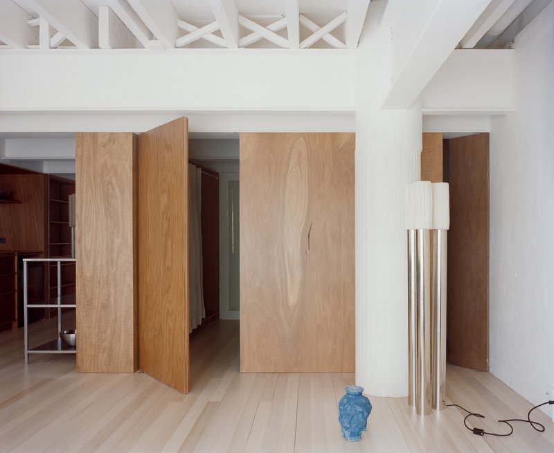 Interior with pivoting wood veneer panels and exposed white-painted roof trusses above pale timber flooring