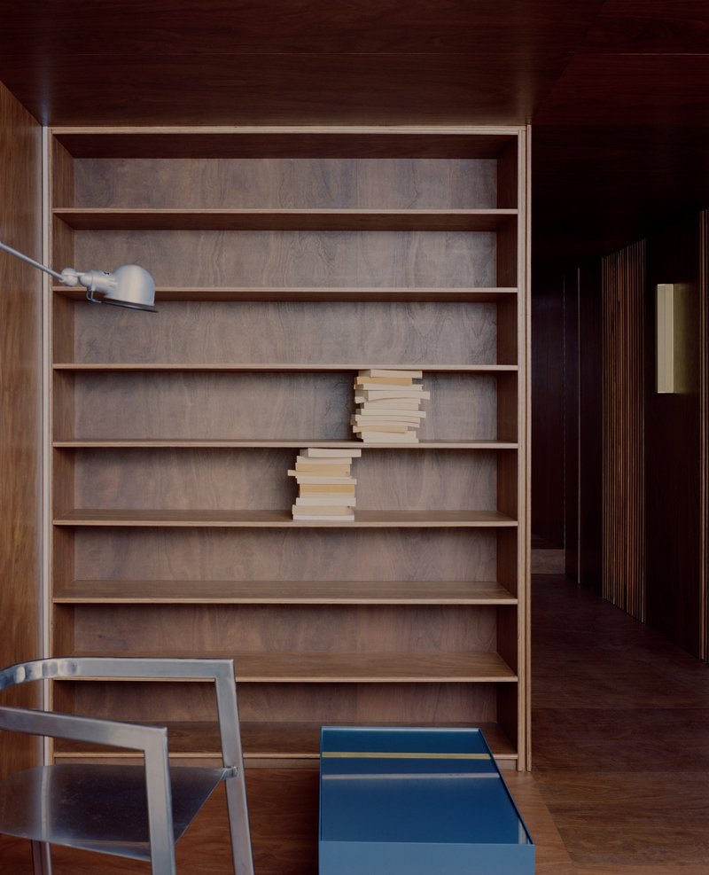 Built-in timber shelving unit with stacked books beside a dark hallway and blue storage box