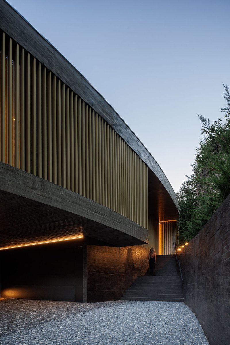 Entrance stairway between concrete retaining walls beneath cantilevered volume with vertical timber louvres at twilight