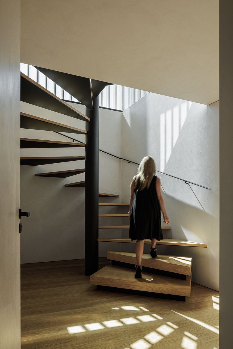 Timber and steel staircase with person ascending beneath a skylight casting geometric shadows across white walls